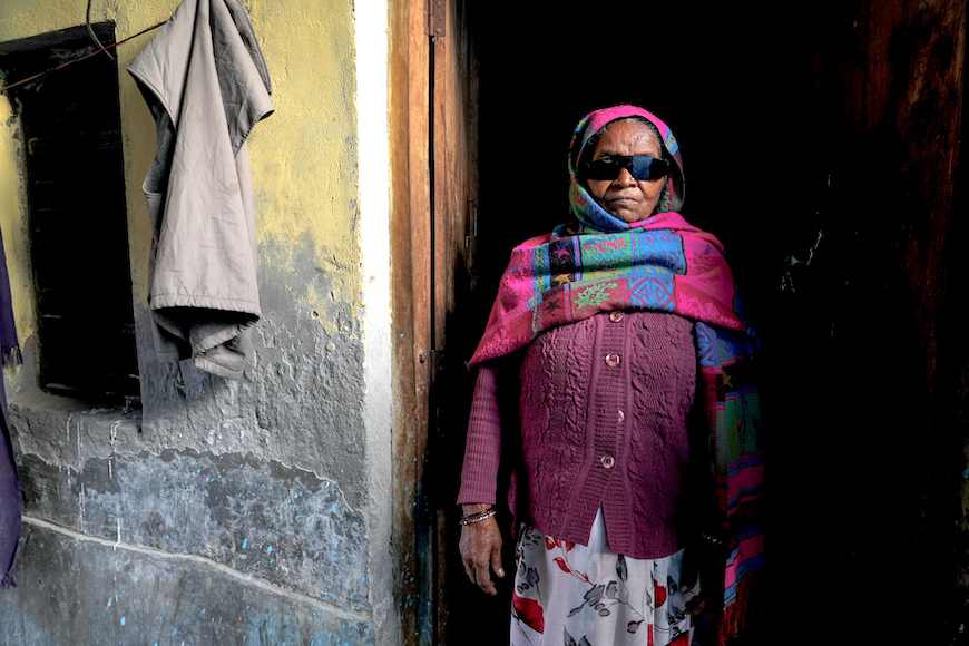 Rupwati, a 65-year-old resident of Uncha Amirpur, stands in a doorway, wearing black glasses.  