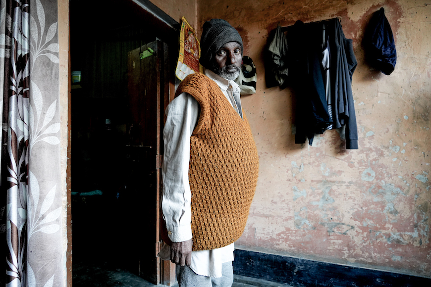 An Indian man, Harinder Singh, a resident of Uncha Amirpur village, stands inside his house, with his head turned right to look at the camera. Clothes hang on wall hooks in the background. Photo: Suhail Bhat 